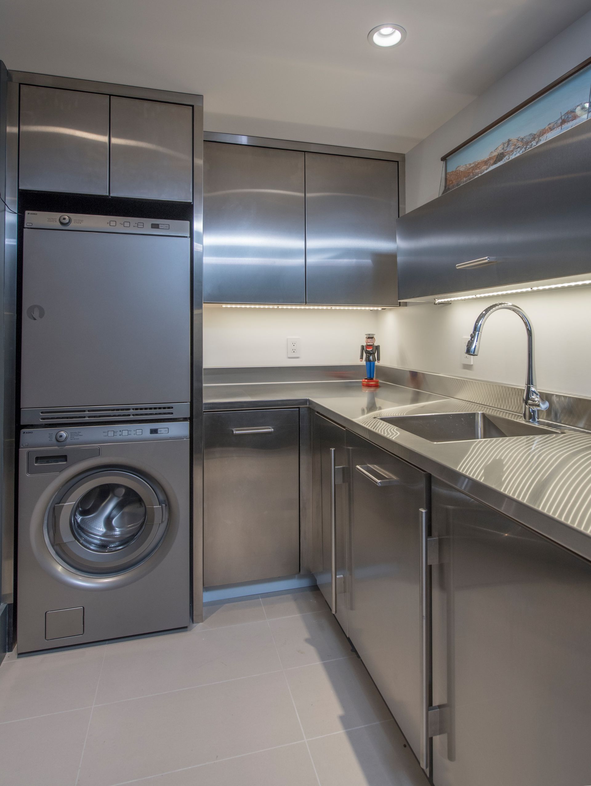 Stainless steel laundry room with washer, dryer, sink, and cabinets; bright lighting and minimalist design.