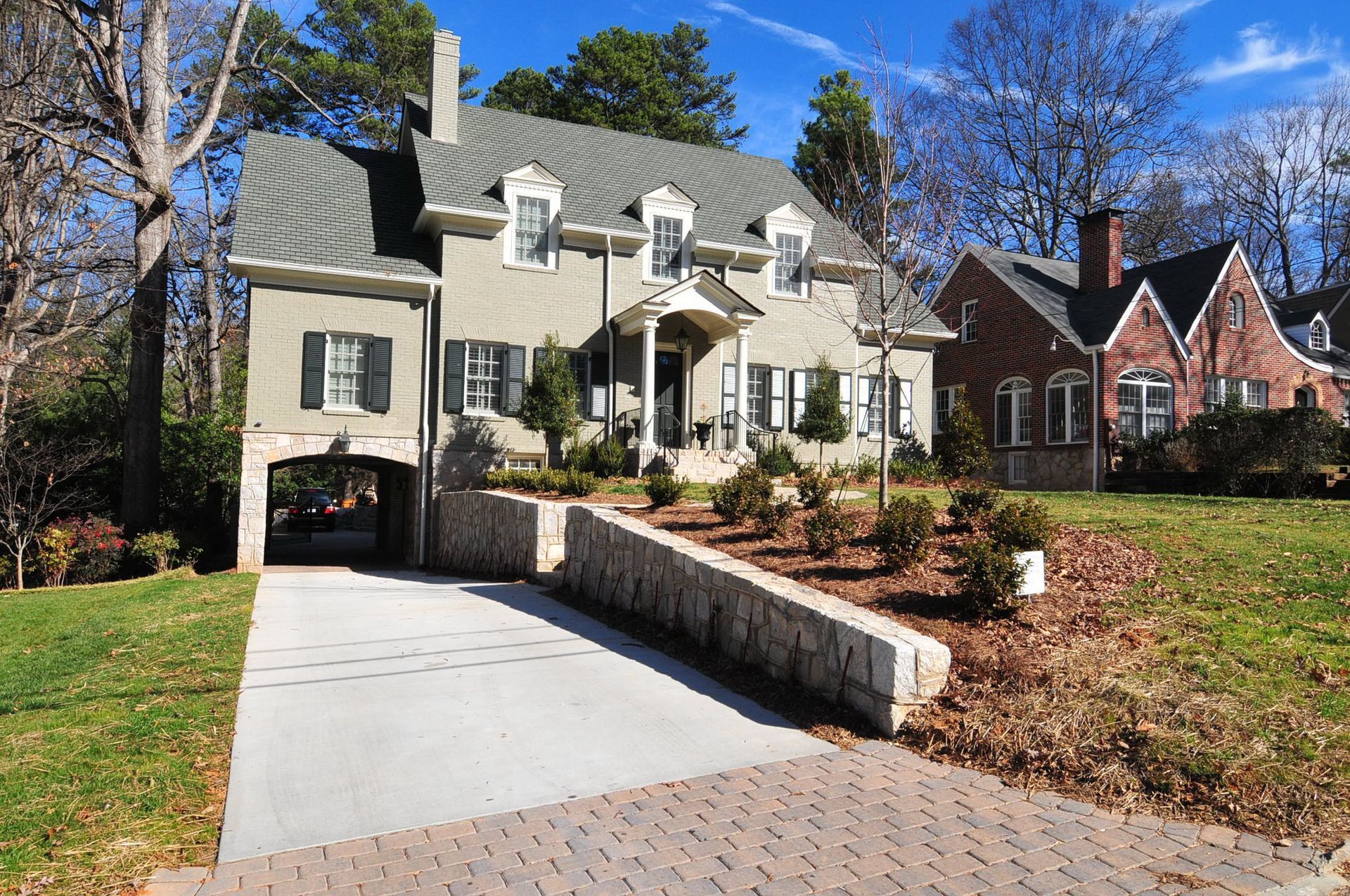 A two-story house with a gray facade, a stone retaining wall, and a brick driveway on a sunny day.