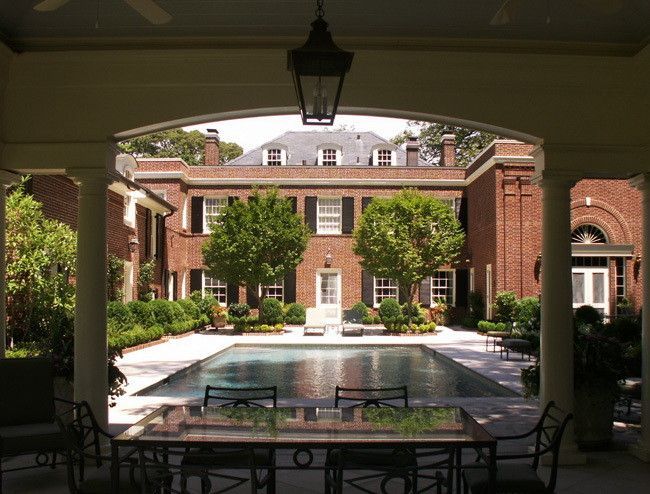 Pool view of a large brick mansion, seen from a shaded patio with a table and chairs.