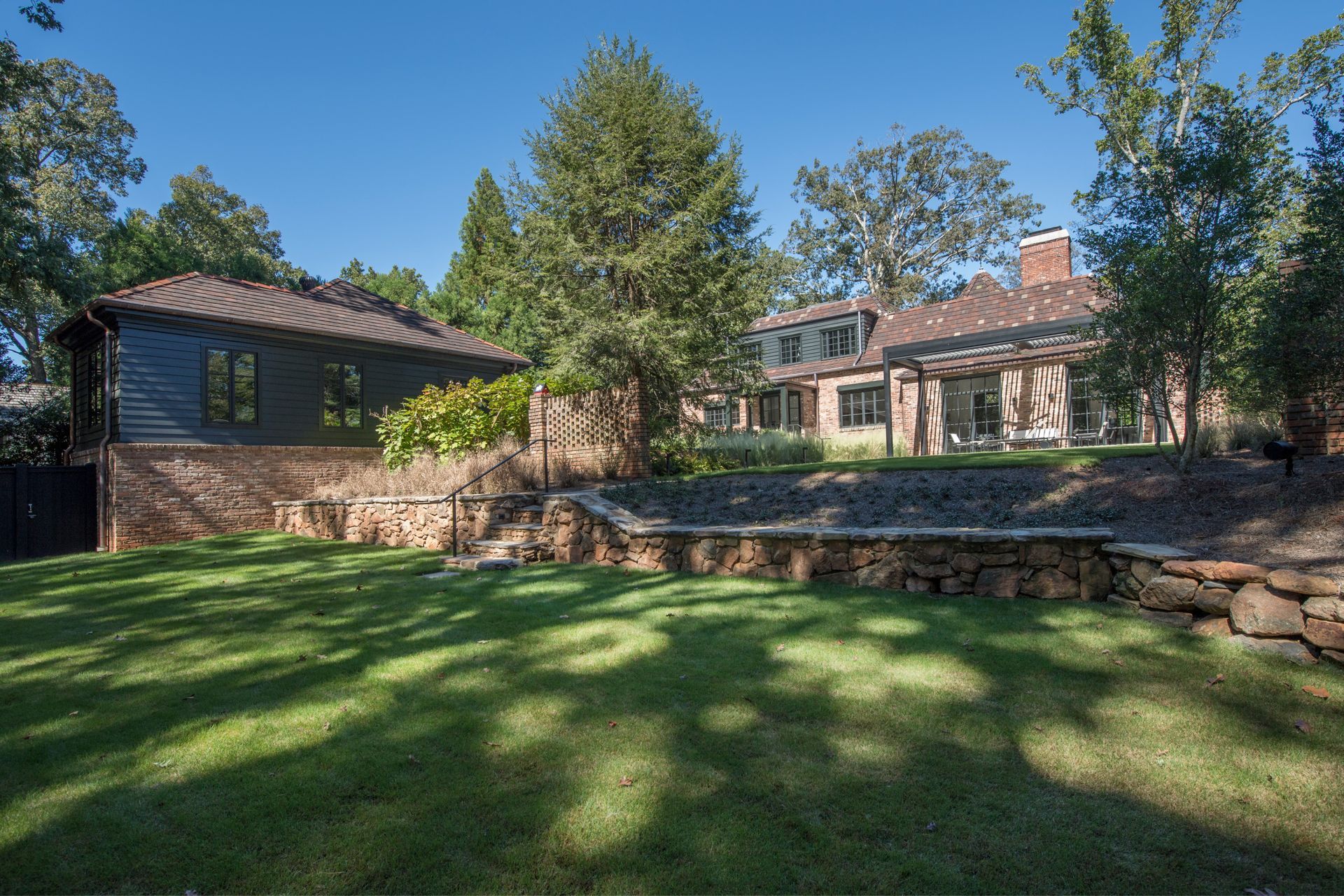 Backyard with a house, lawn, and stone retaining walls under a blue sky.