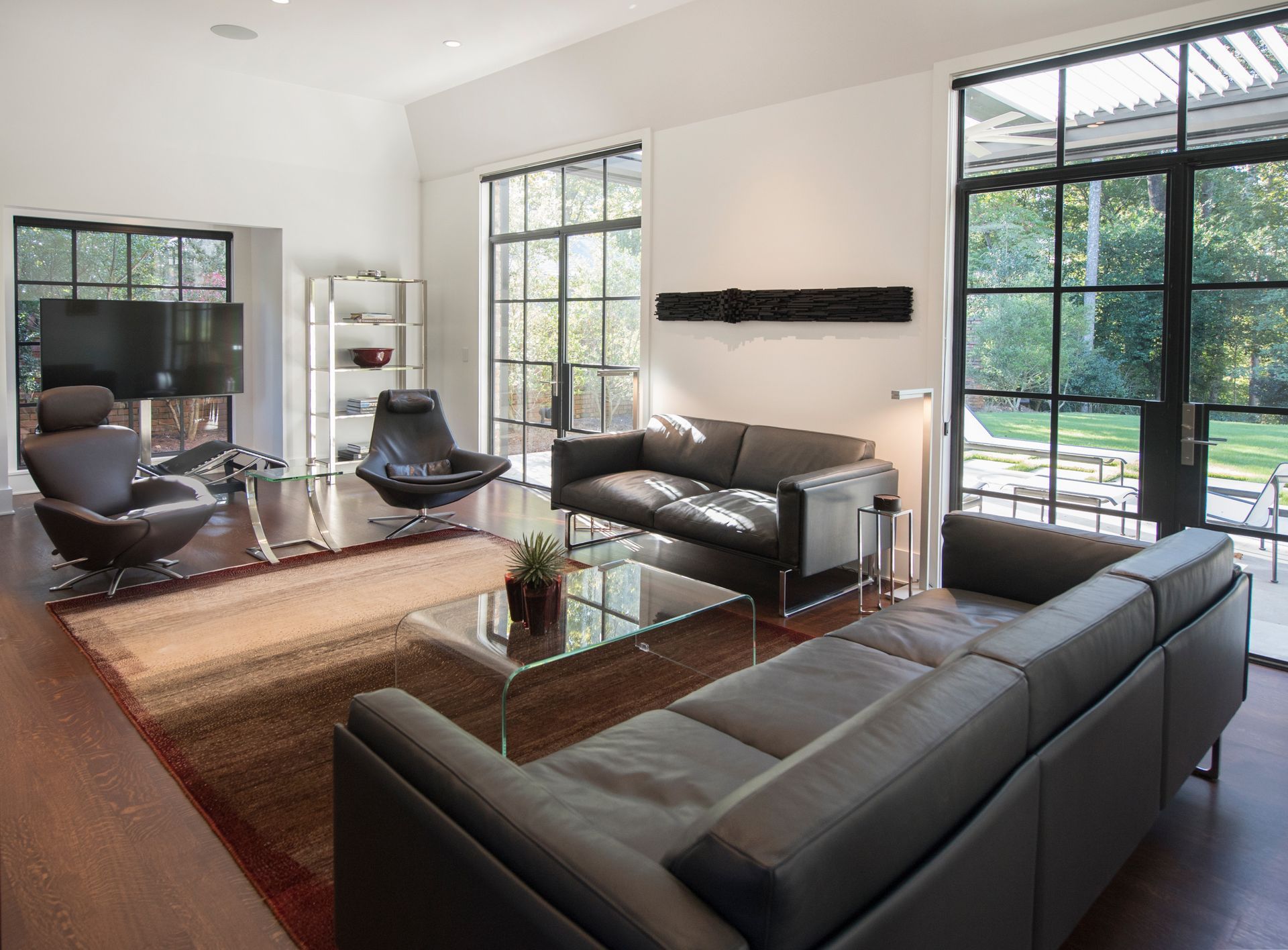 Modern living room with black leather sofas, large windows, and a view of a yard.