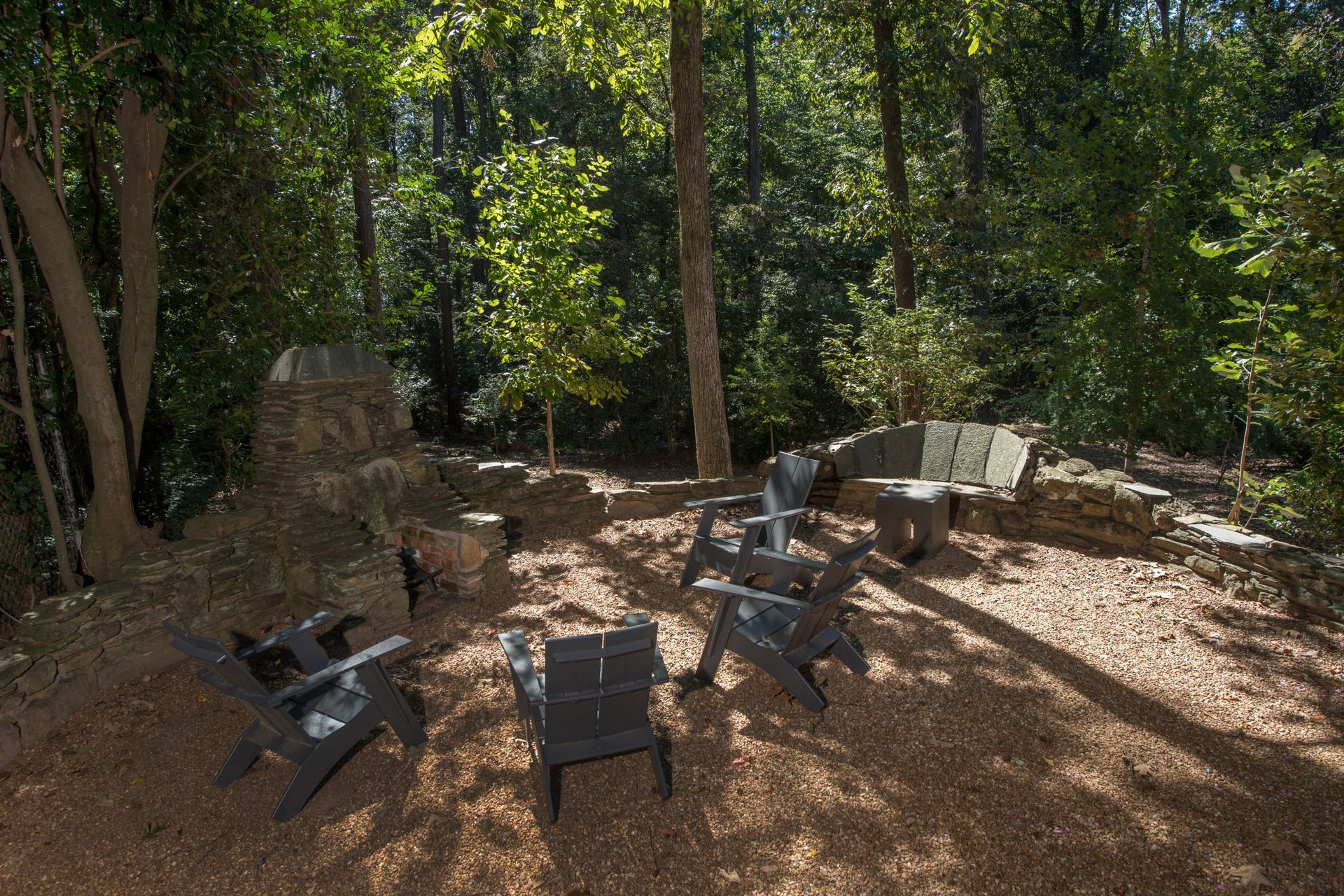 Outdoor seating area with dark chairs around a stone fireplace in a wooded setting.