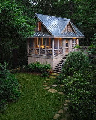 Quaint wooden cabin with screened porch nestled in lush green forest. Stone path leads to the steps.