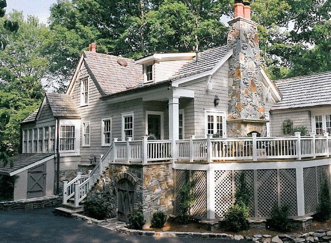 Gray cottage with stone chimney and deck, surrounded by trees.