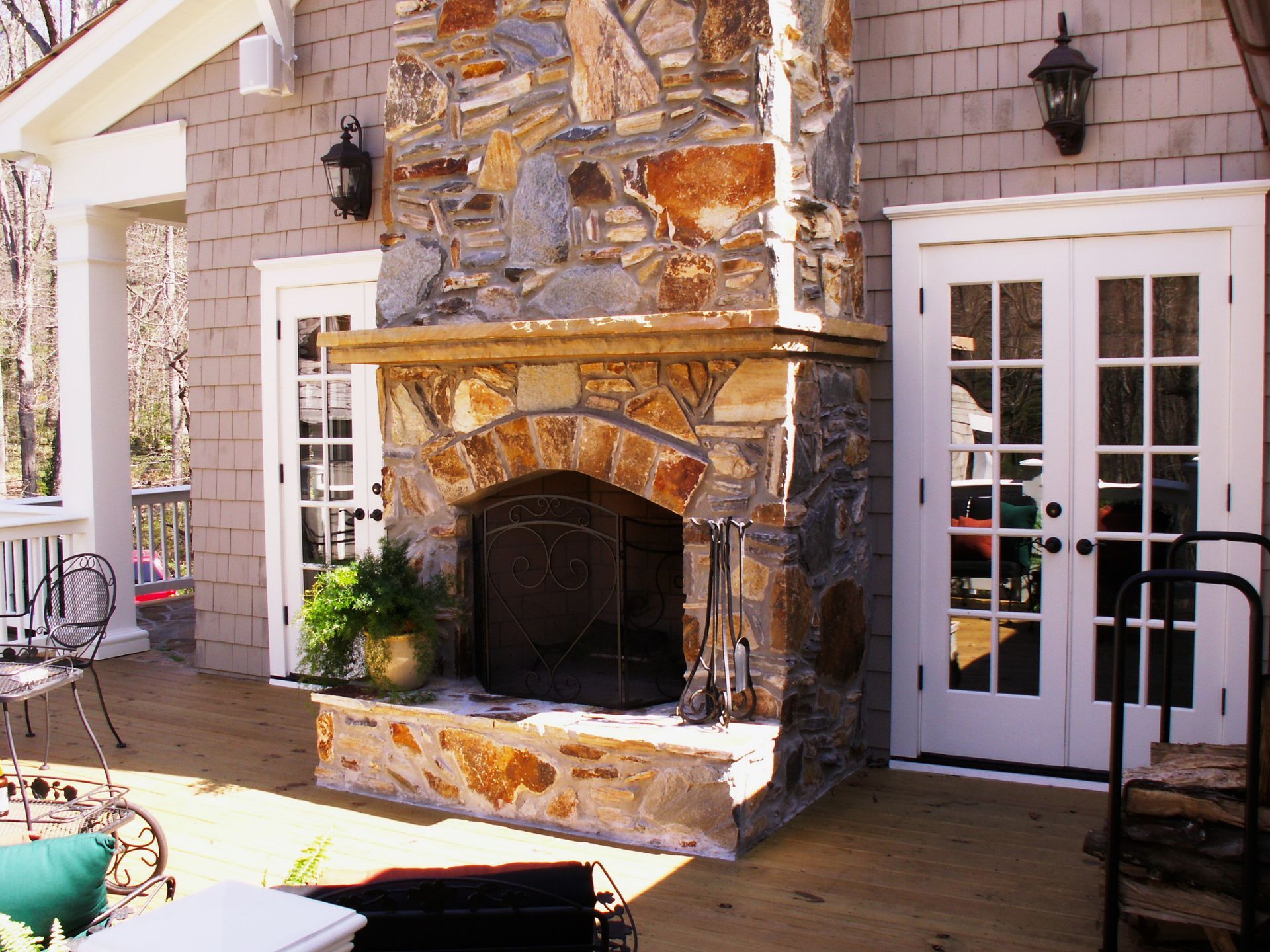 Outdoor stone fireplace with French doors, flanked by sconces, on a wooden deck.