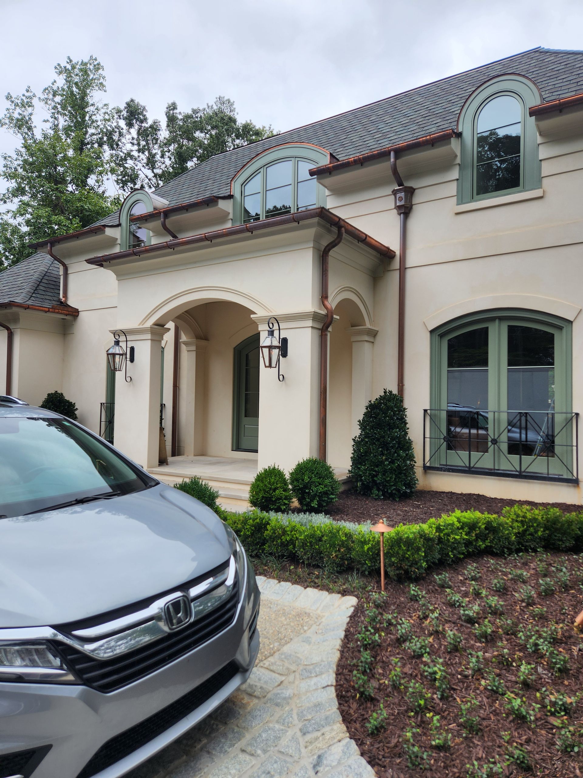 A beige stucco house with a gray car parked in front. Green windows, dark trim, and a stone walkway.
