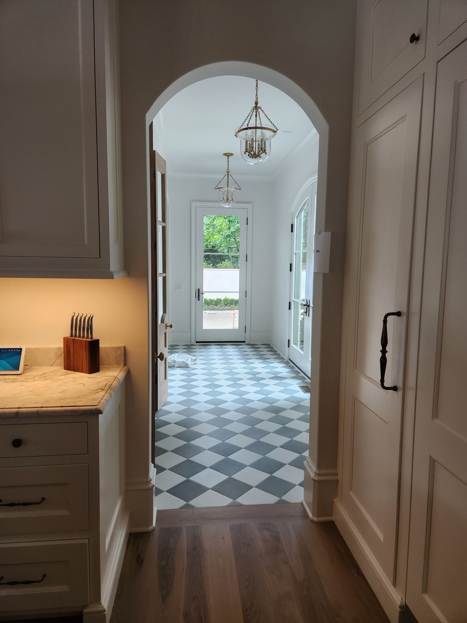 Hallway with checkered tile floor leading to a door.  Arched doorway, white walls, and light fixtures.