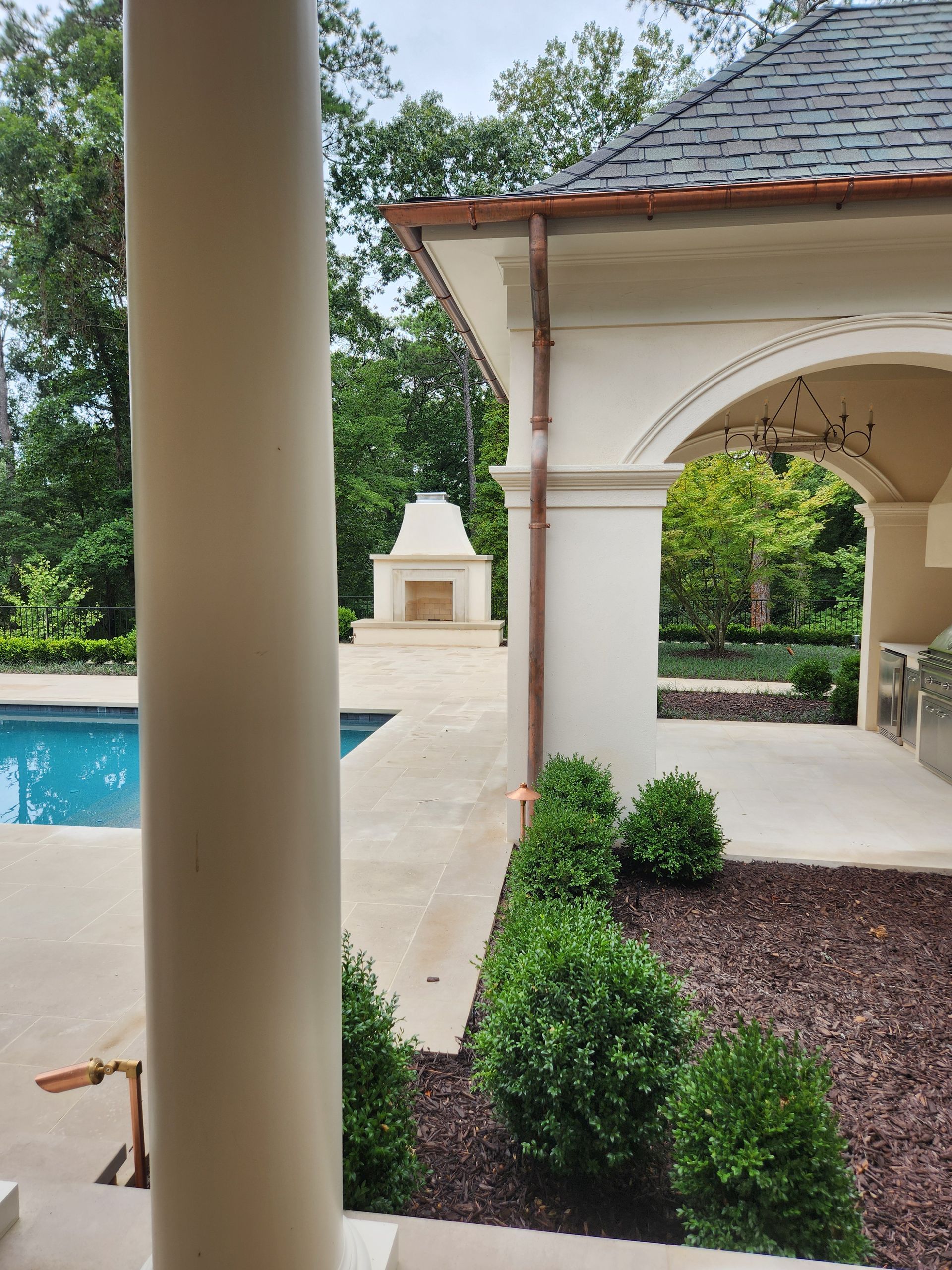 Poolside view: outdoor kitchen, fireplace, pool, beige architecture, green shrubbery, trees in background.