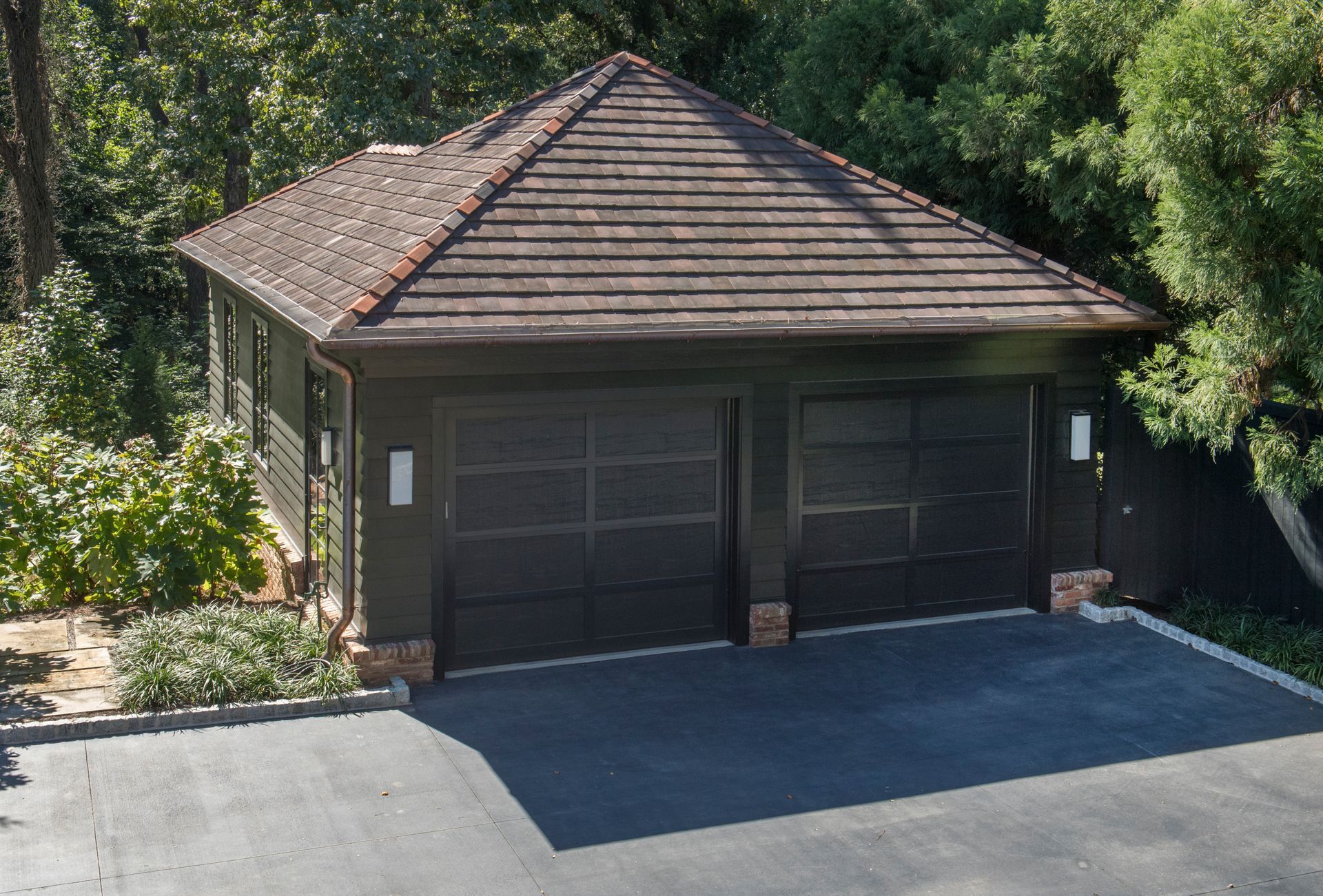 Dark garage with two doors and a shingled roof, surrounded by trees and asphalt.