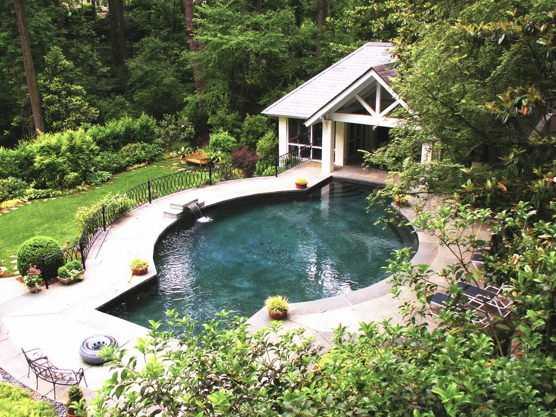 Pool with grotto-like cabana surrounded by lush greenery. Stone pool deck with landscaping.