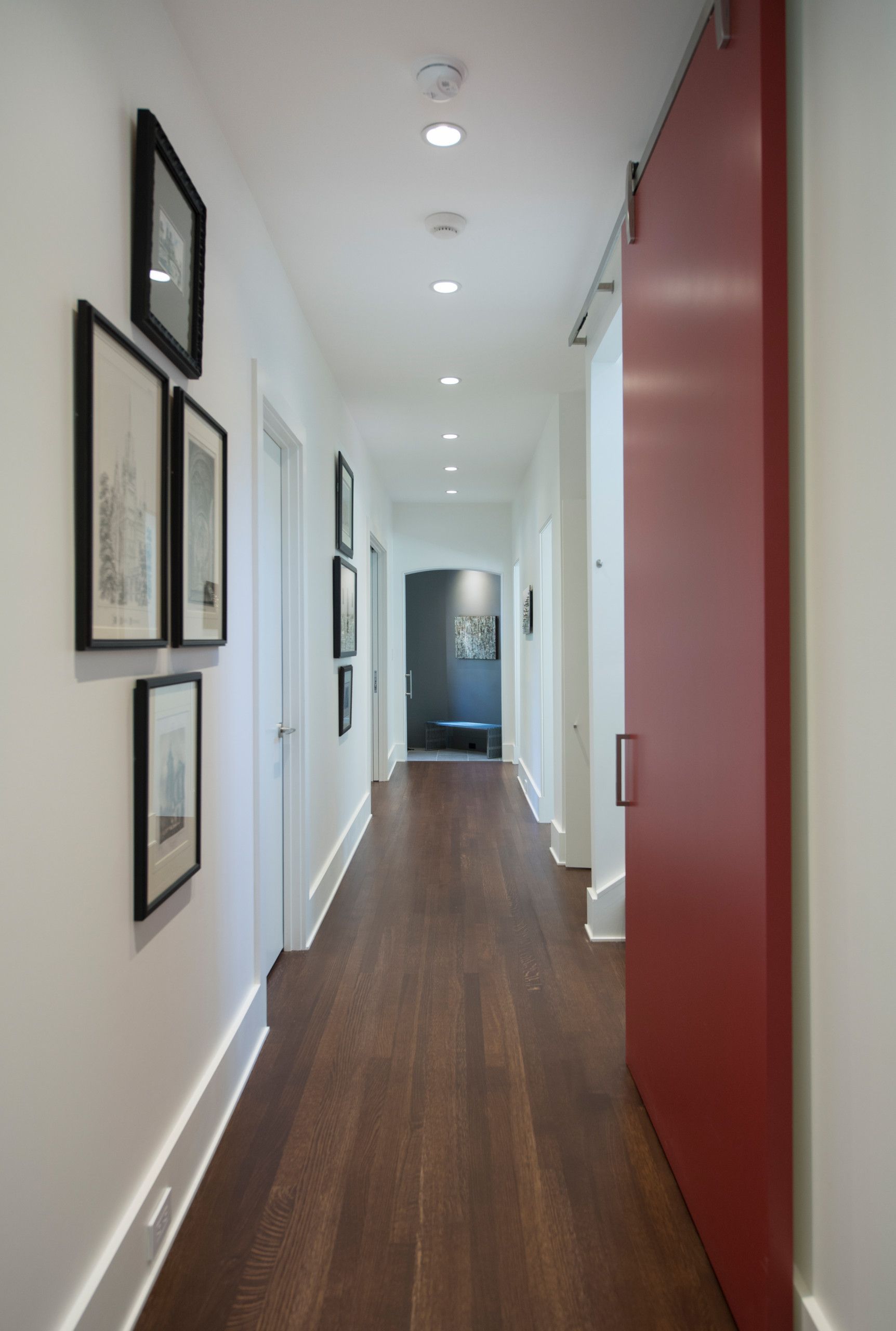 Long hallway with dark wood floor, white walls, artwork, and a red sliding door.