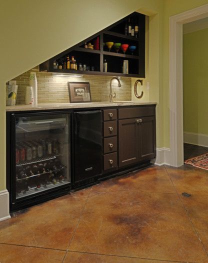 Small bar under stairs with a mini-fridge, sink, dark brown cabinets, and shelves with bottles and glasses; brown flooring.