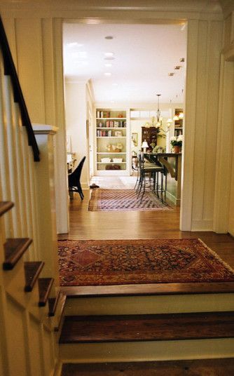 Wooden staircase leads to hallway with rug, kitchen, and bookshelves.