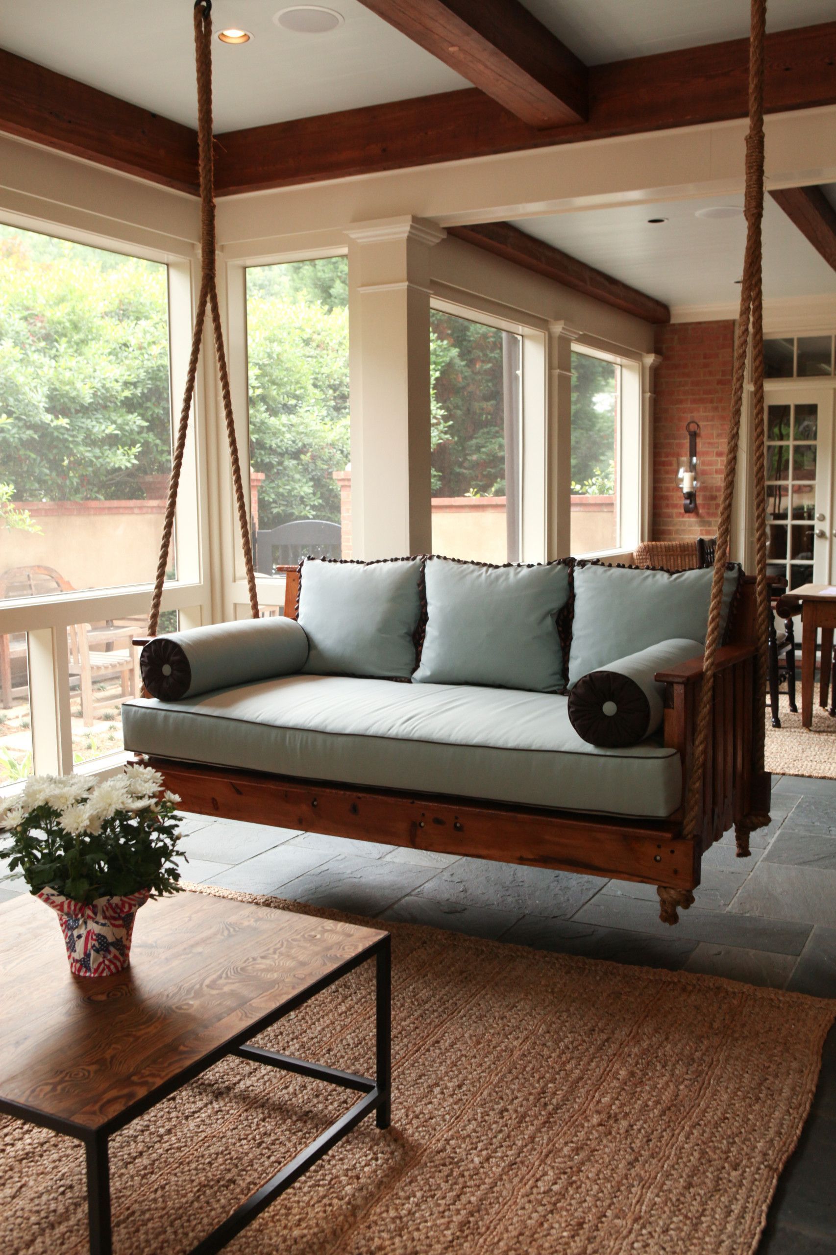 Outdoor porch with hanging swing, light blue cushions, wooden frame. Brown rug, coffee table, brick wall.
