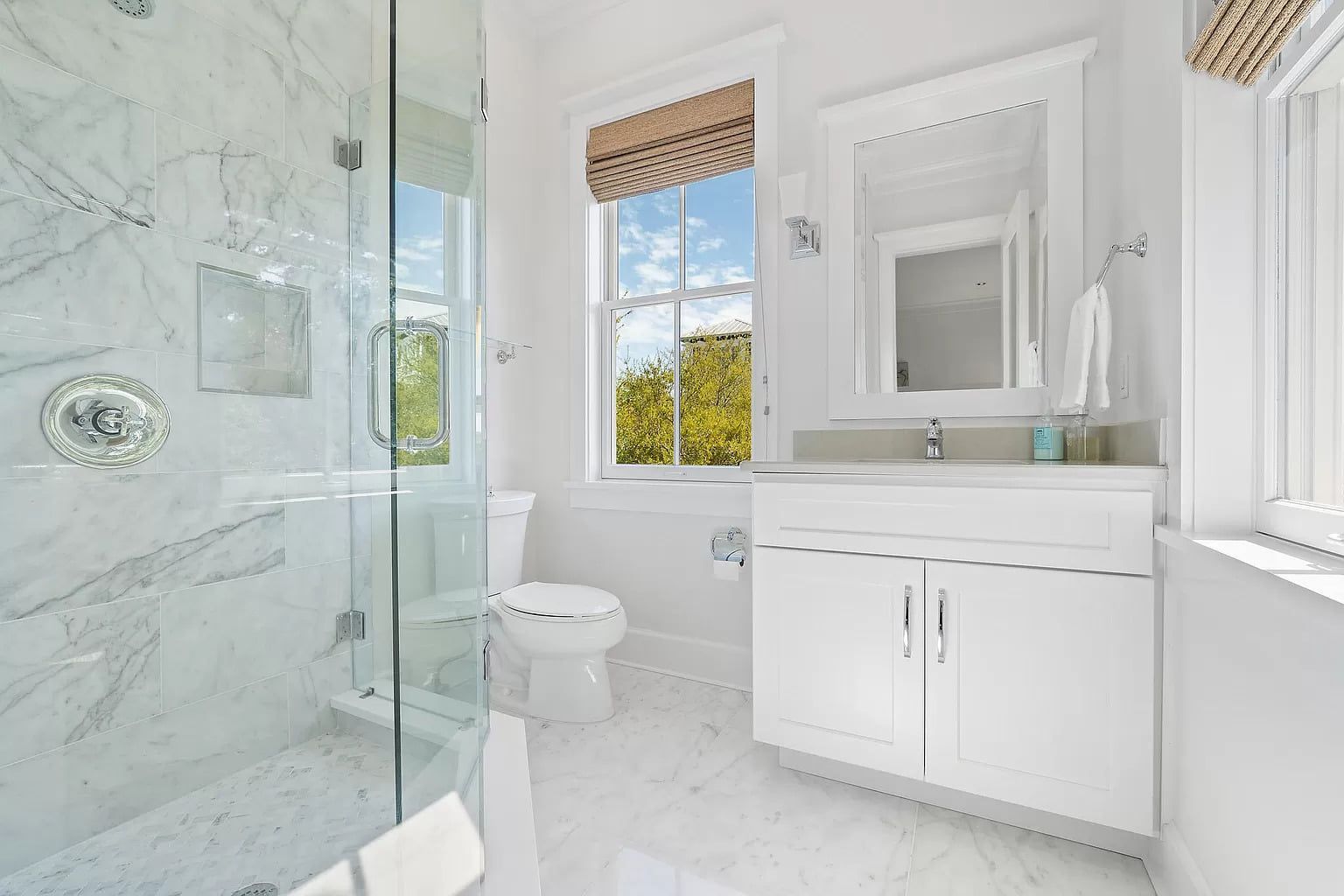 Bright, white bathroom with a glass shower, toilet, sink, and window with a view.