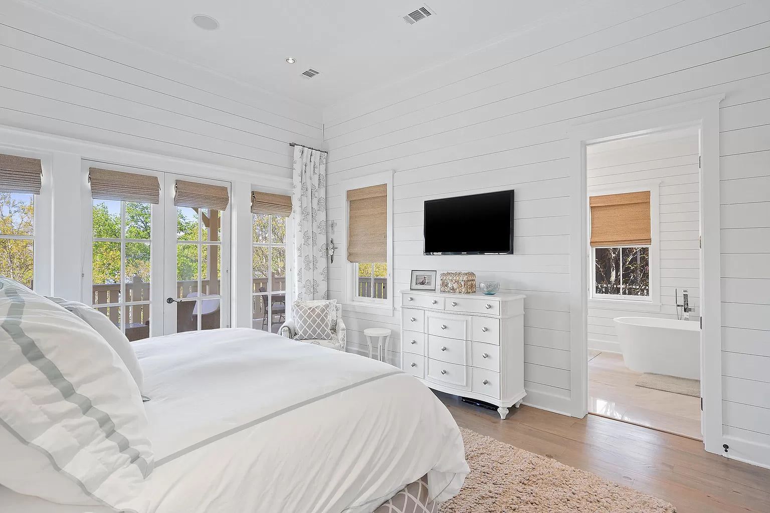 Bright white bedroom with bed, dresser, TV, and doorway to a bathroom with a tub.