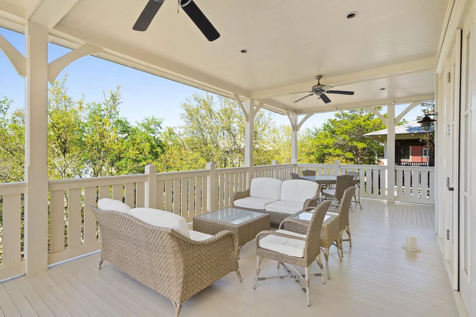 Covered outdoor patio with white railing, wicker furniture, ceiling fans, and trees in the background.