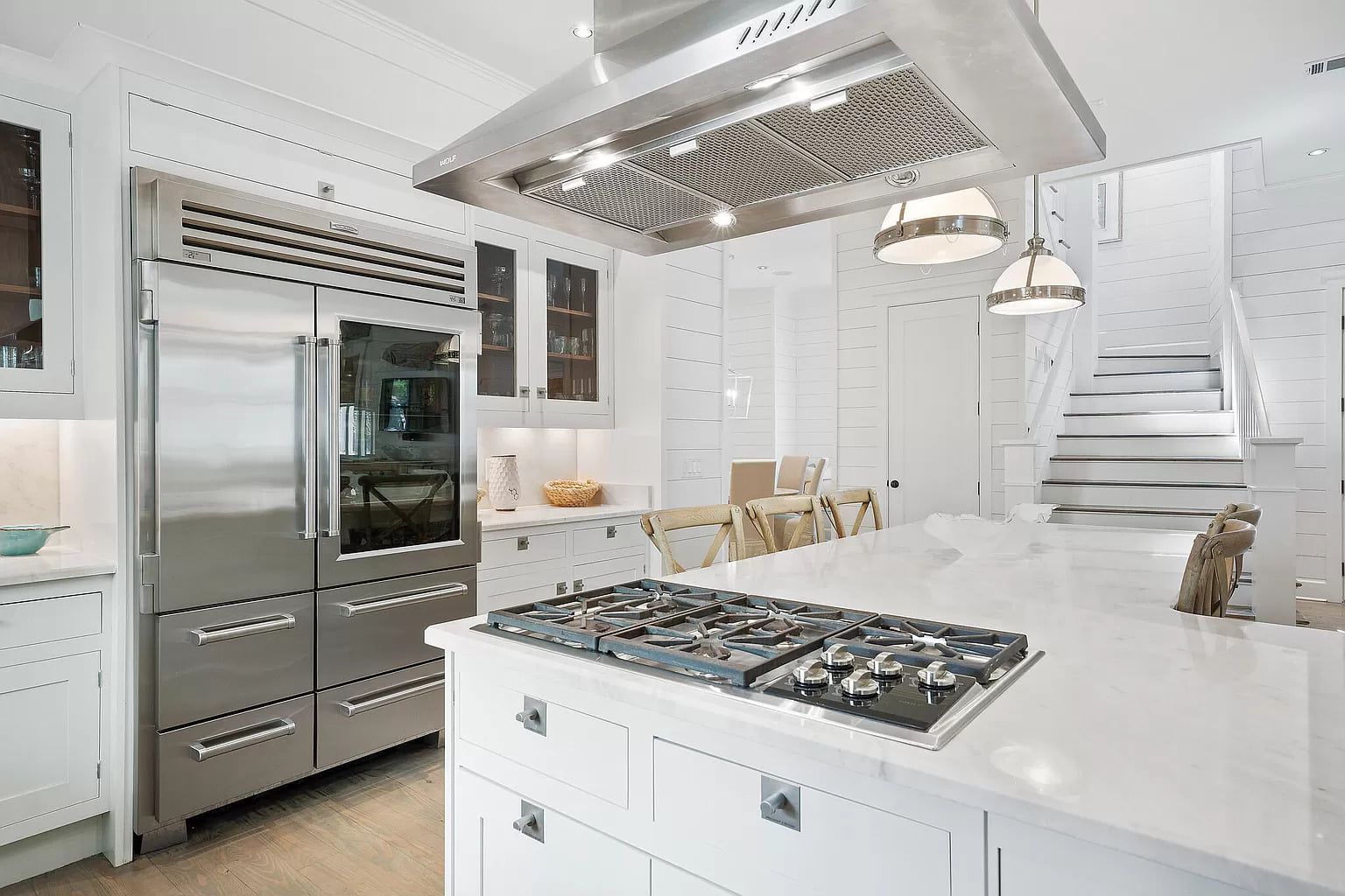 Modern white kitchen with stainless steel appliances, island with cooktop, and cat near stairs.