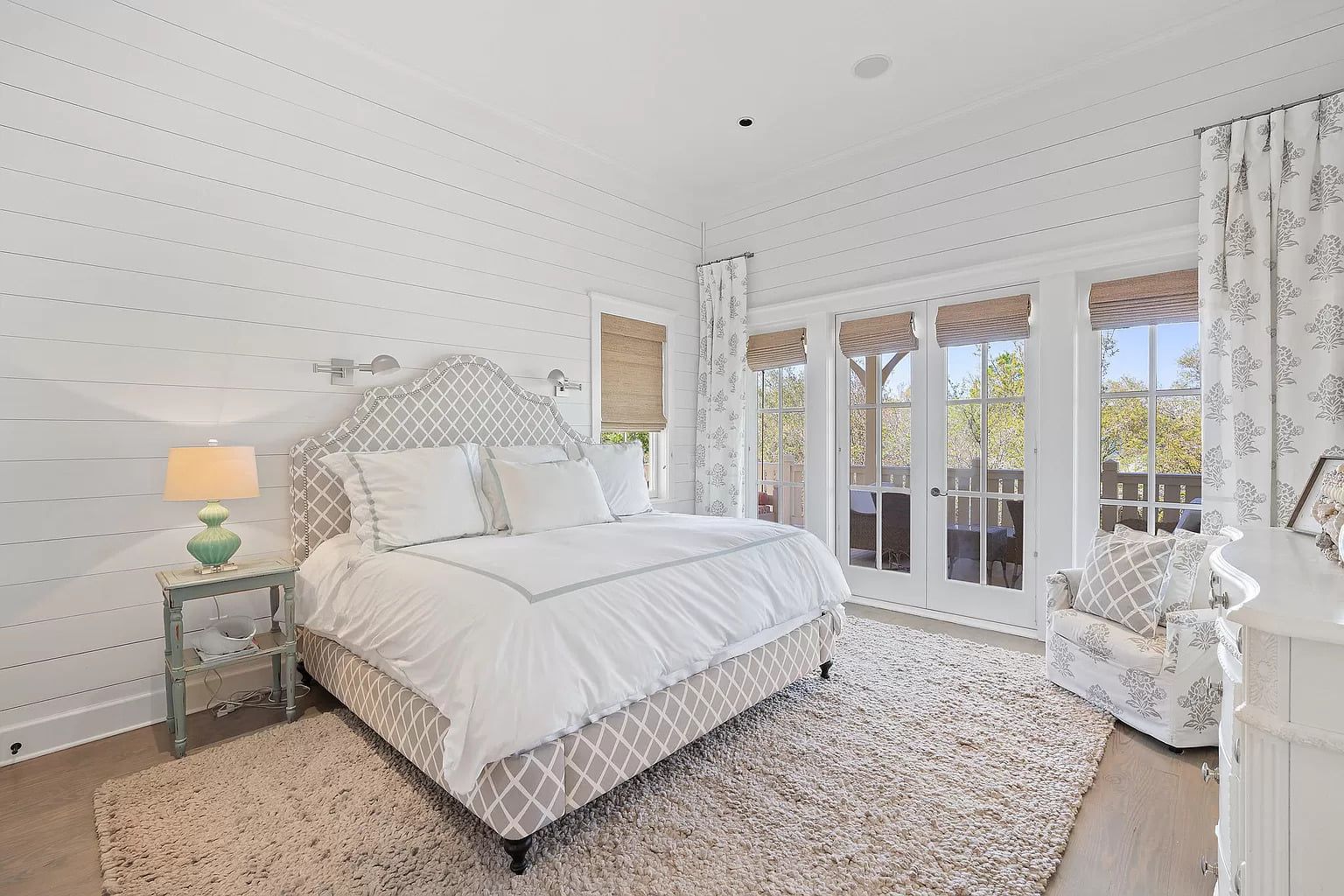 Bright bedroom with white and beige decor; bed with patterned headboard, doors to a balcony.