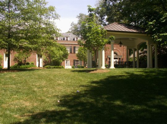 Brick building with white columns and gazebo in a grassy yard, trees surrounding.
