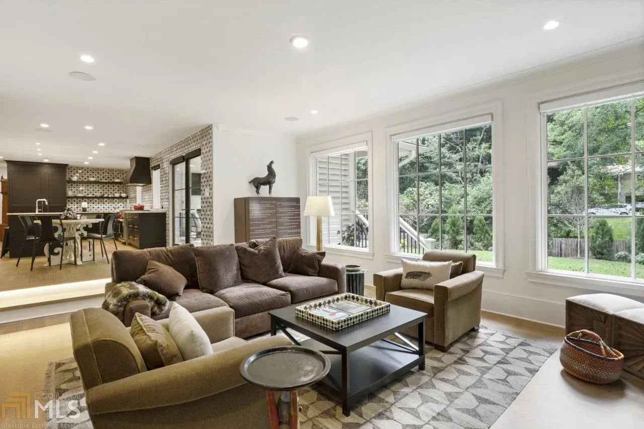 Living room with brown sofa, armchairs, coffee table, and large windows overlooking a yard.
