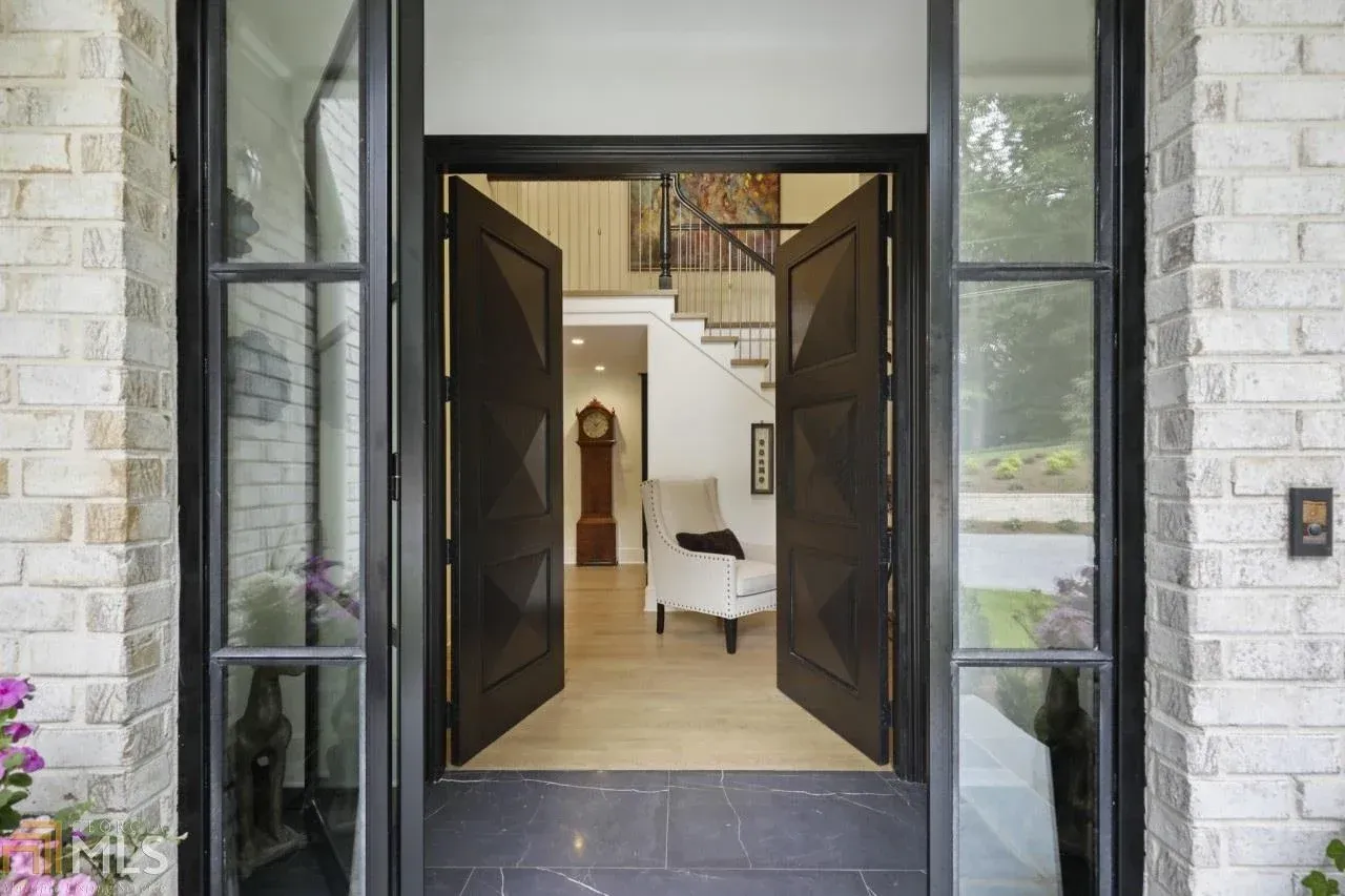 Open black double doors reveal interior with staircase, grandfather clock, and armchair. Brick exterior.