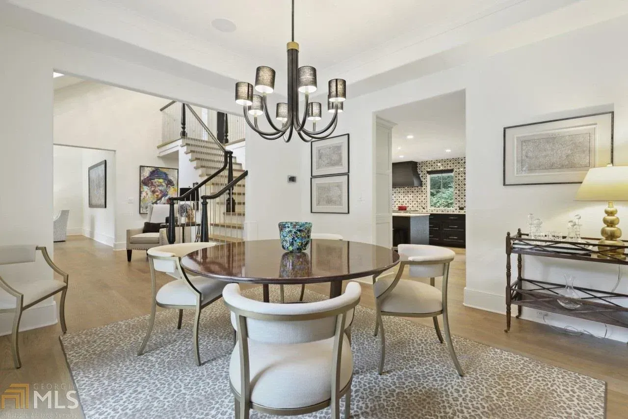 Elegant dining room with round table, chandelier, and open staircase. Light walls, carpet, and chairs.