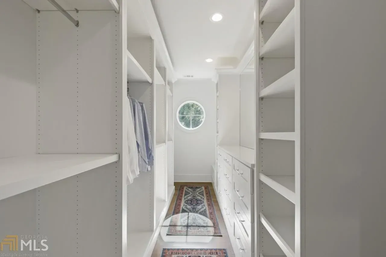 White walk-in closet with shelves and drawers. Round window at the end, blue and beige rug.