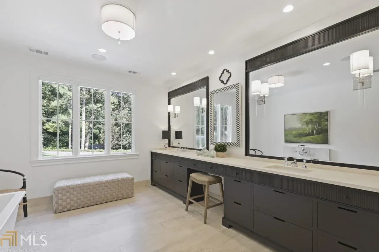 Spacious bathroom with dark cabinets, long mirrors, white walls, and a window looking out to greenery.