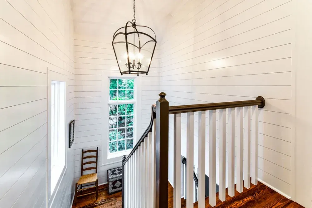 Stairwell with white shiplap walls, wood railing, and a lantern-style light fixture. A window overlooks a green tree.