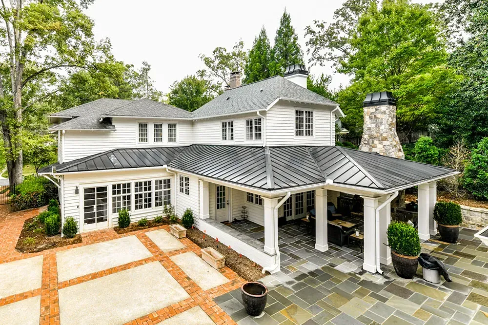 White two-story house with black roof, open-air patio, brick and stone paving, surrounded by green trees.