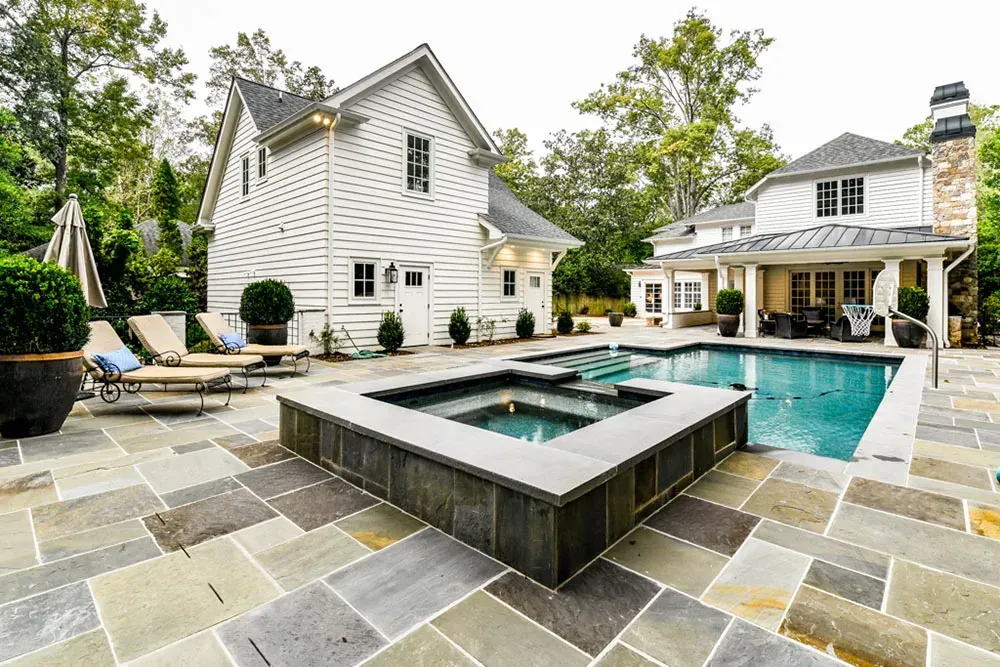 Backyard with pool and spa, two-story white house, stone patio, lounge chairs, trees in background.