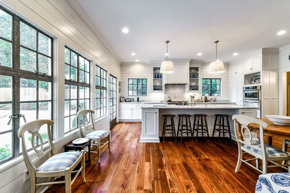 Bright kitchen with hardwood floors, white cabinets, and large windows with black trim; chairs sit by the windows.