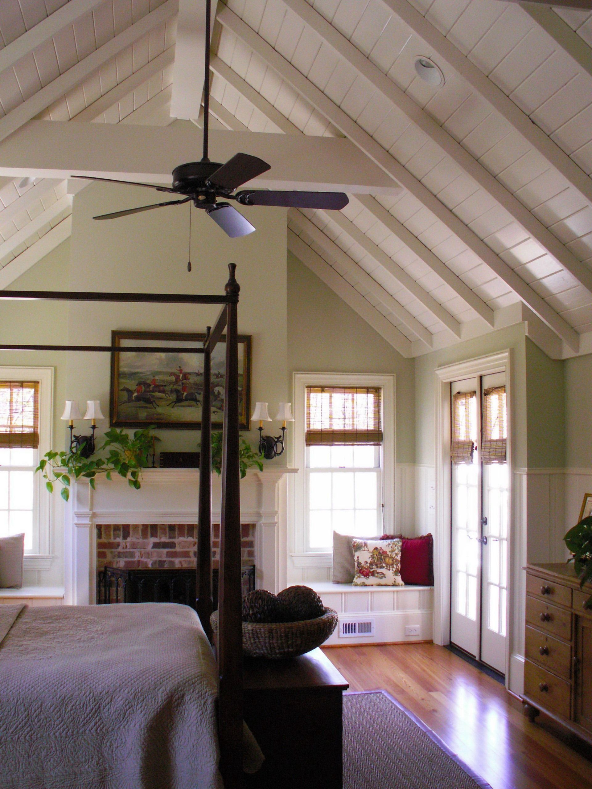 Bedroom with high, beamed ceiling, brick fireplace, four-poster bed, and windows with woven shades; light green walls.