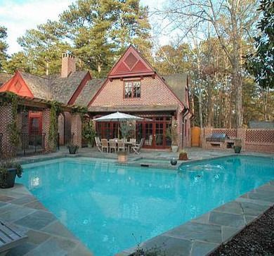 A luxurious brick house with a pool, patio, and outdoor dining area in a wooded setting.