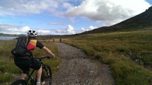 Person on mountain bike riding on a gravel path through grassy terrain, a mountain and lake in the background.