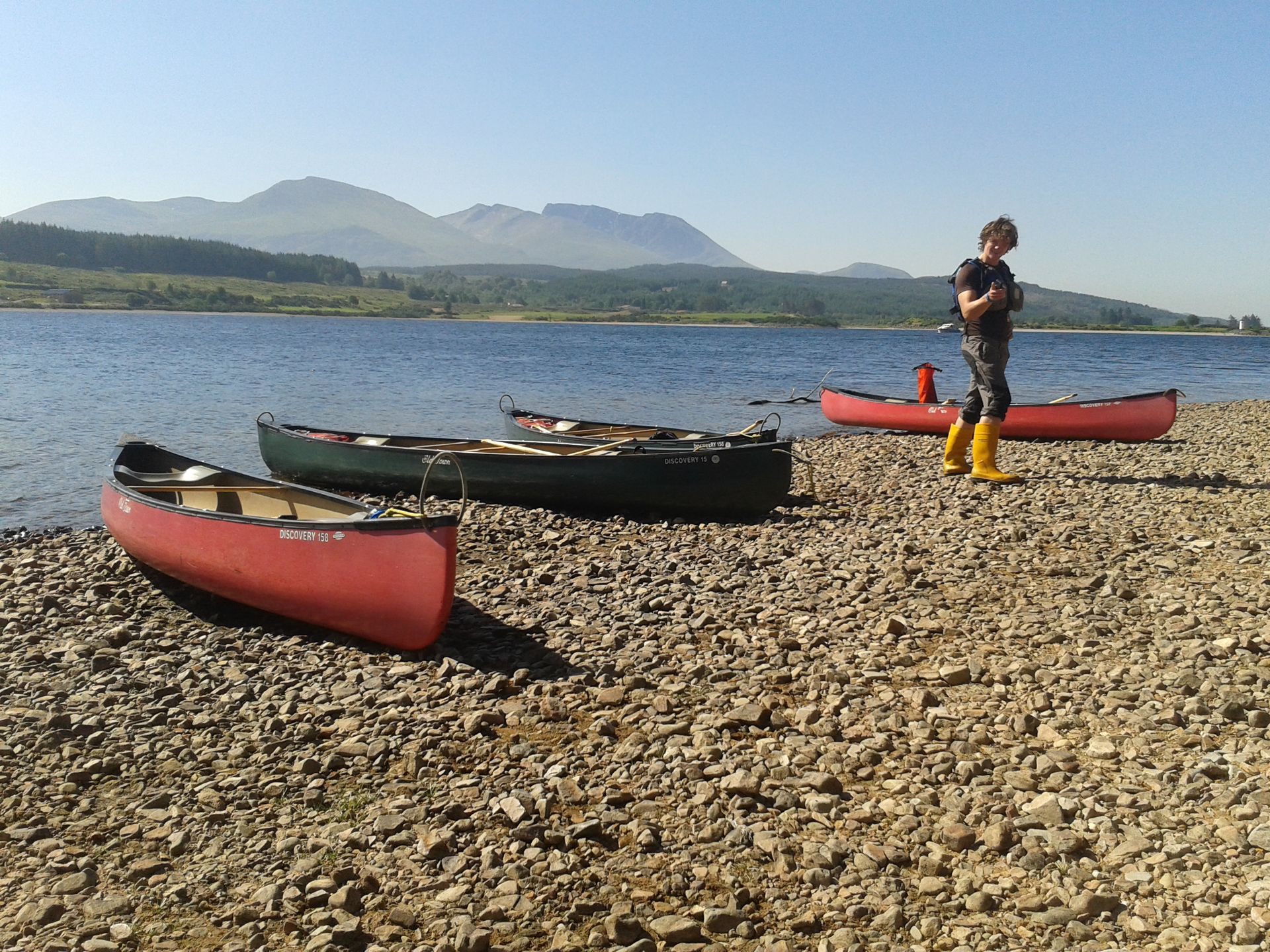Four canoes on a rocky shore, a person in yellow boots stands nearby, mountains and water in the background.