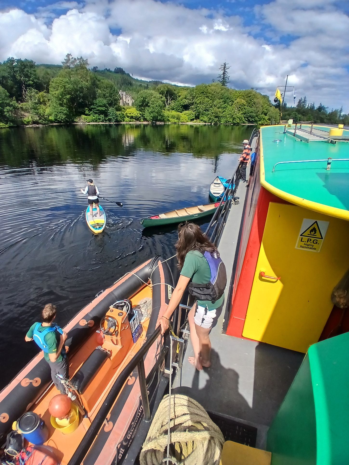 People by a colorful boat on a waterway; some standup paddleboard, canoe. Trees and building in background.