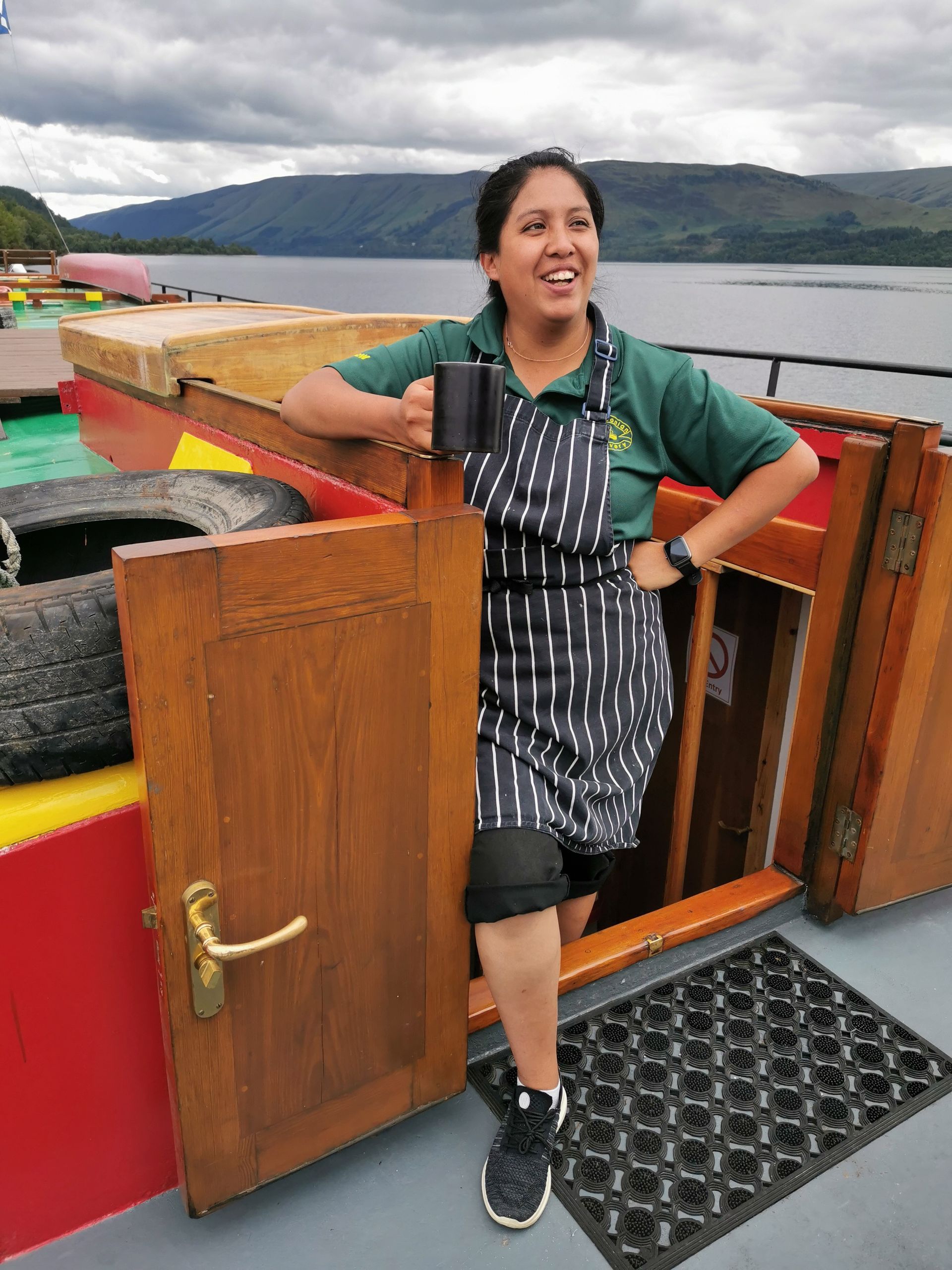 Woman on a boat, holding a mug and smiling, wearing an apron, in front of a doorway, scenic view in background.