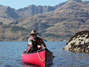 Man in red canoe with dog on water, mountains in background.