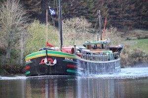 A boat named Inga, with a black hull and green accents, sails on a river. Scottish flag flies.