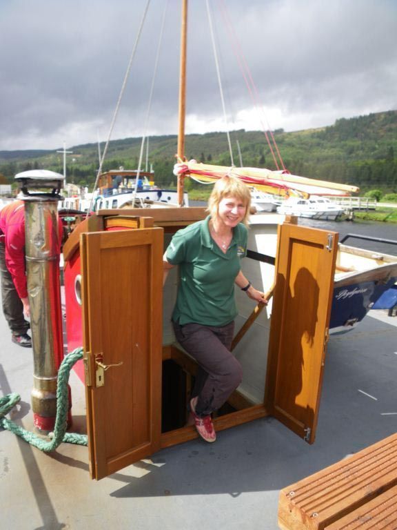 Woman stepping out of a wooden boat cabin onto the deck, wearing a green shirt, looking at the camera.