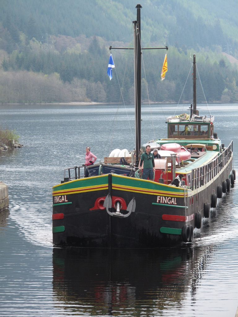 Black barge on water with mast and flags; green and red accents, man at the helm.