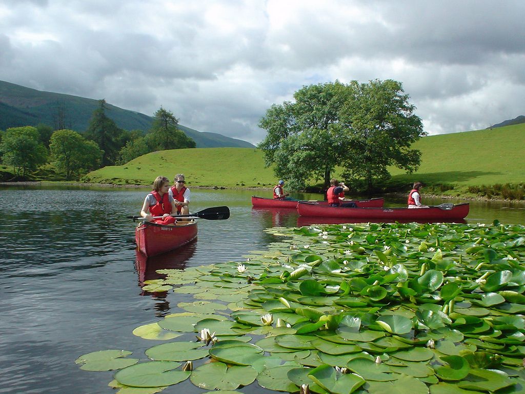 People canoeing on a lake, with lily pads in the foreground and green hills in the background under a cloudy sky.