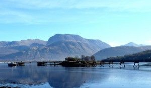 Blue lake scene with bridge, small islands, and mountains under a clear sky.