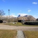 Grassy area with lamp post, small buildings, and snow-capped mountains under a blue sky.