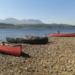 Red canoes on a rocky shore, overlooking a blue lake and distant mountains under a clear sky.