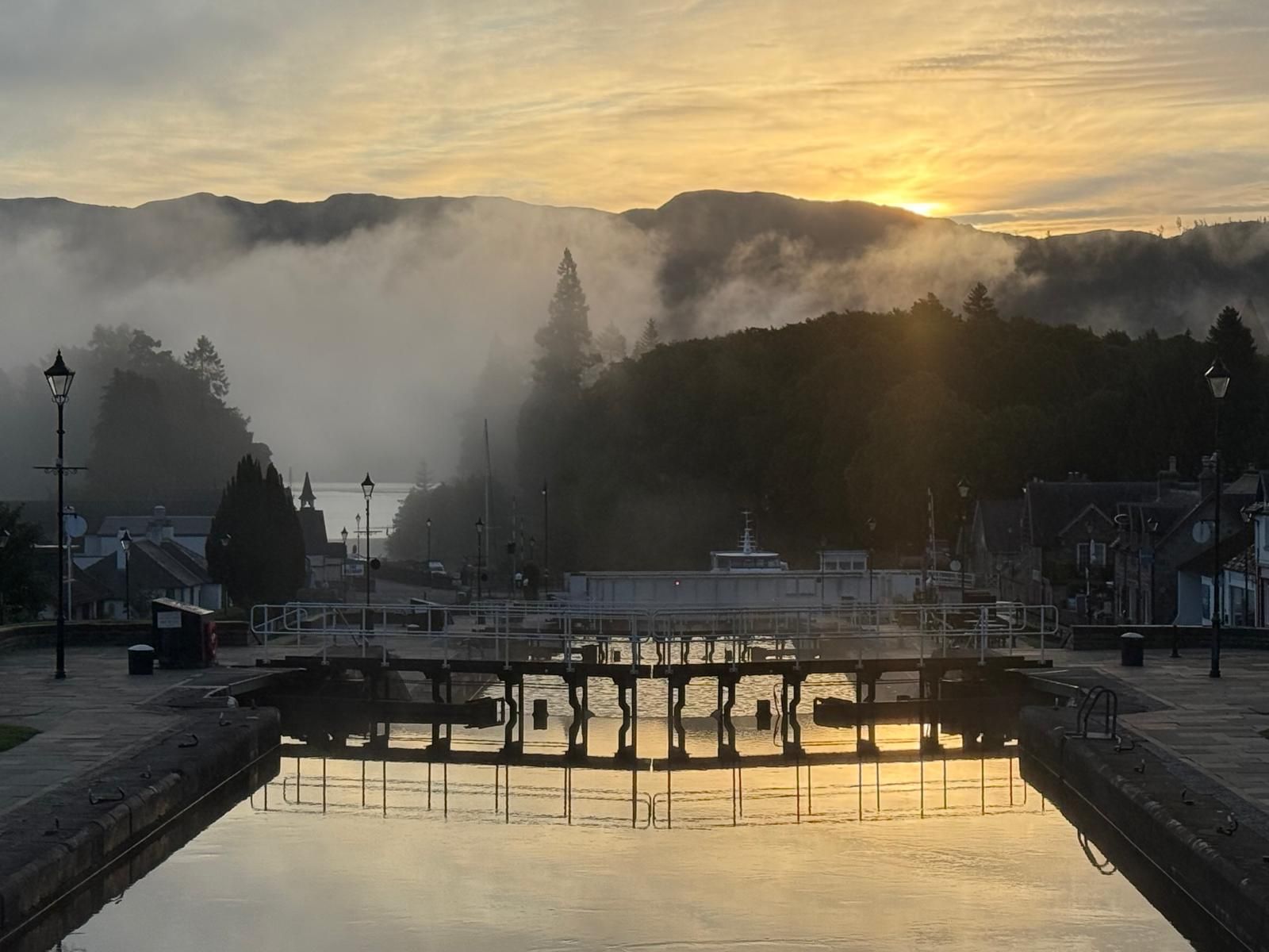 Sunrise over a canal with lock gates. Fog blankets the hills.
