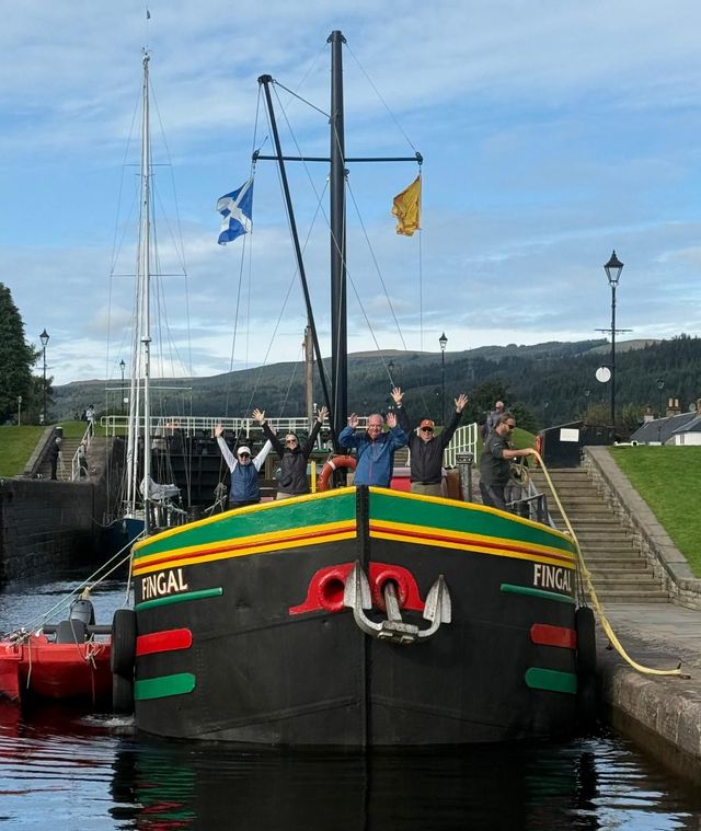 A group of people on a black boat, with arms raised, in a canal lock, with Scottish flags and green hills.