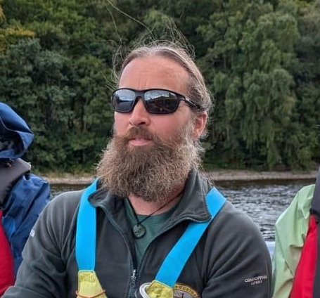 Man with beard and sunglasses wearing a teal medal, in front of water and trees.